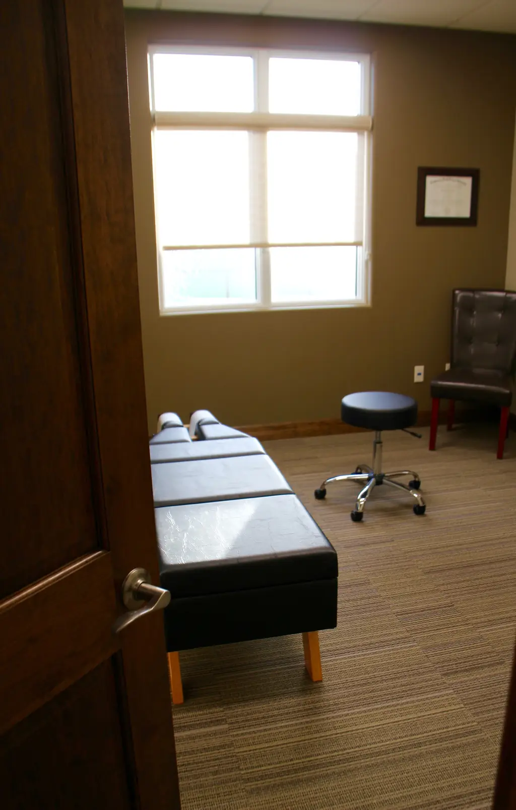 Doorway view into a treatment room at Alpha Chiropractic with bench seating and a framed diploma